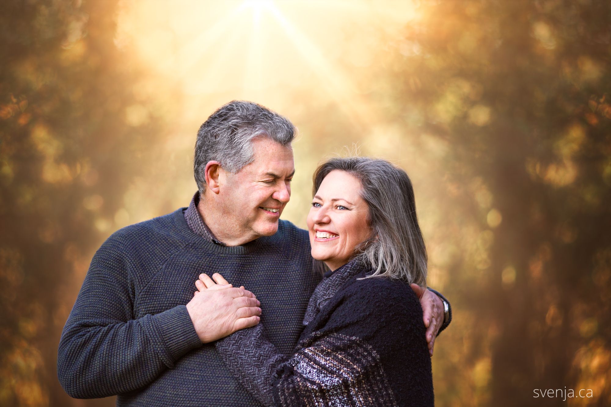 husband and wife share a smile while he holds her in front of trees with sun beams behind them
